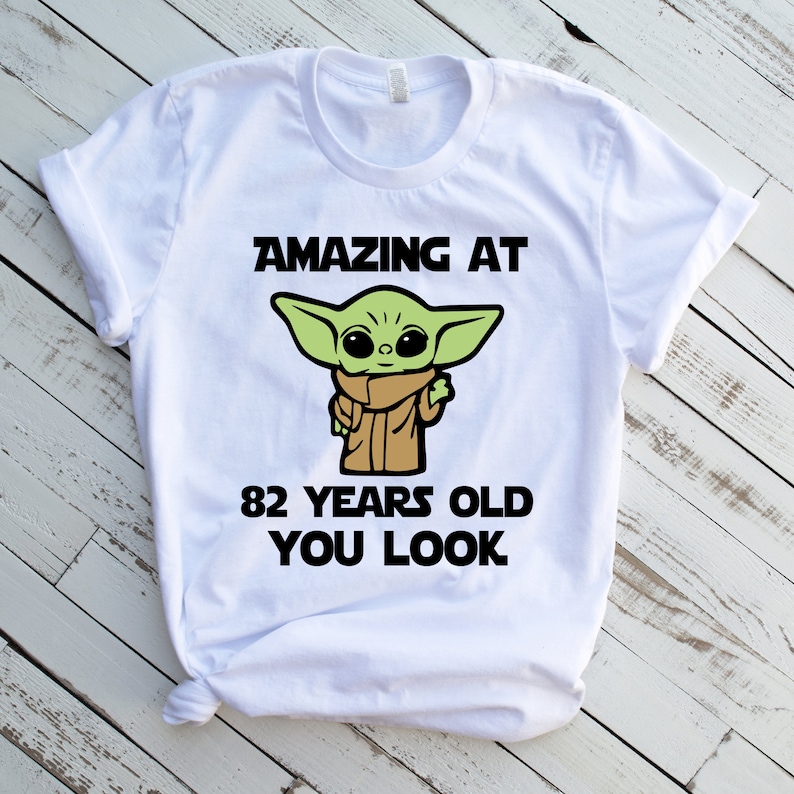 May include: White t-shirt with a green cartoon character and the text "AMAZING AT 82 YEARS OLD YOU LOOK".