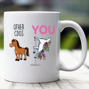May include: White ceramic coffee mug with a pink "YOU" printed on the right side. A unicorn is depicted on the right side of the mug, with a rainbow mane and tail, holding onto a pole. On the left side of the mug, a brown horse is depicted. The text "OTHER CDOS" is printed above the horse.