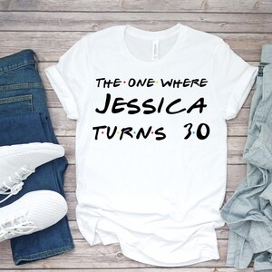 May include: White t-shirt with the text "THE ONE WHERE JESSICA TURNS 30" in black, red, and yellow. The shirt is displayed on a wooden surface with blue jeans and white sneakers.