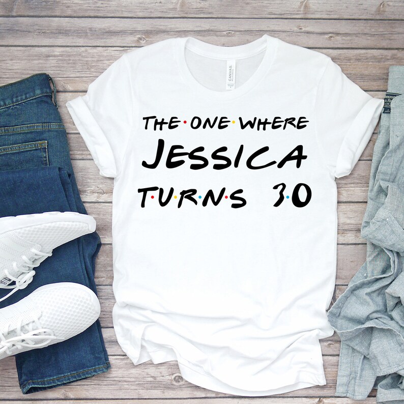 May include: White t-shirt with the text "THE ONE WHERE JESSICA TURNS 30" in black, red, and yellow. The shirt is displayed on a wooden surface with blue jeans and white sneakers.