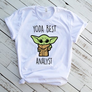 May include: White t-shirt featuring a cartoon image of Grogu, also known as Baby Yoda, with the text "YODA BEST ANALYST" in black. The character is green with large eyes and is wearing a brown cloak.