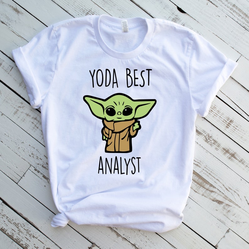 May include: White t-shirt featuring a cartoon image of Grogu, also known as Baby Yoda, with the text "YODA BEST ANALYST" in black. The character is green with large eyes and is wearing a brown cloak.