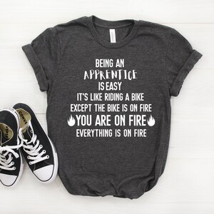 May include: A dark grey t-shirt with white text that reads "Being an Apprentice is Easy. It's like riding a bike except the bike is on fire. You are on fire. Everything is on fire."