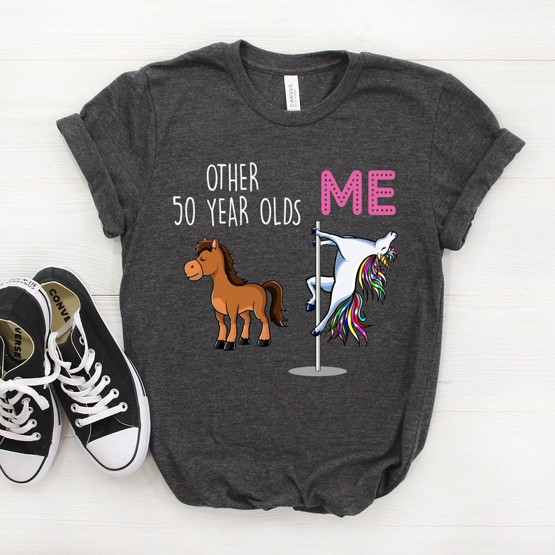 Pode incluir: Uma camiseta cinza escura com o texto "Other 50 year olds" e "Me" impresso nela. A palavra "Me" est&aacute; em rosa e est&aacute; ao lado de um unic&oacute;rnio de desenho animado fazendo uma dan&ccedil;a de barra. A palavra "Other" est&aacute; ao lado de um cavalo marrom de desenho animado.