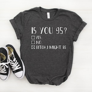 May include: Dark gray t-shirt with the text "IS YOU 95?" and check boxes for "YES", "NO", and "BITCH, I MIGHT BE". Black canvas sneakers with white soles and laces are in the foreground.
