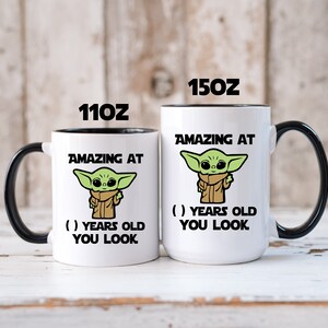 May include: Two white coffee mugs with black rims. The mugs feature a cartoon image of a green alien character with the text "Amazing at ( ) years old you look". The mugs are available in two sizes: 11 oz and 15 oz.