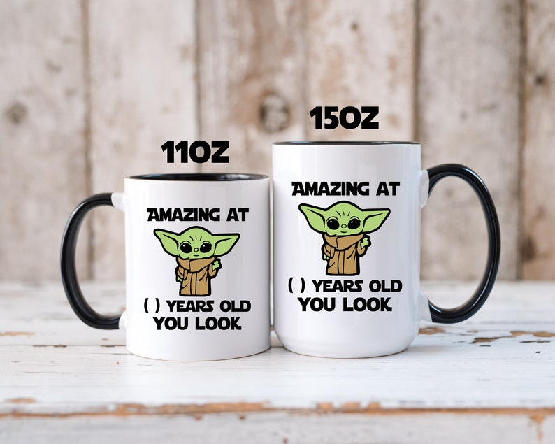 May include: Two white coffee mugs with black rims. The mugs feature a cartoon image of a green alien character with the text "Amazing at ( ) years old you look". The mugs are available in two sizes: 11 oz and 15 oz.