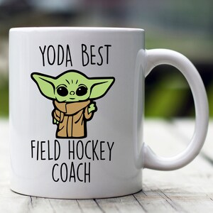 Może przedstawiać: Biały kubek ceramiczny z zieloną postacią z kresk&oacute;wki i tekstem "YODA BEST FIELD HOCKEY COACH".