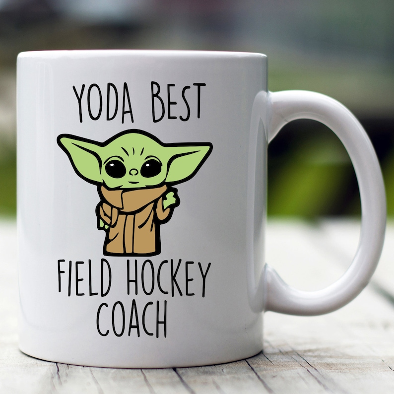 Może przedstawiać: Biały kubek ceramiczny z zieloną postacią z kresk&oacute;wki i tekstem "YODA BEST FIELD HOCKEY COACH".