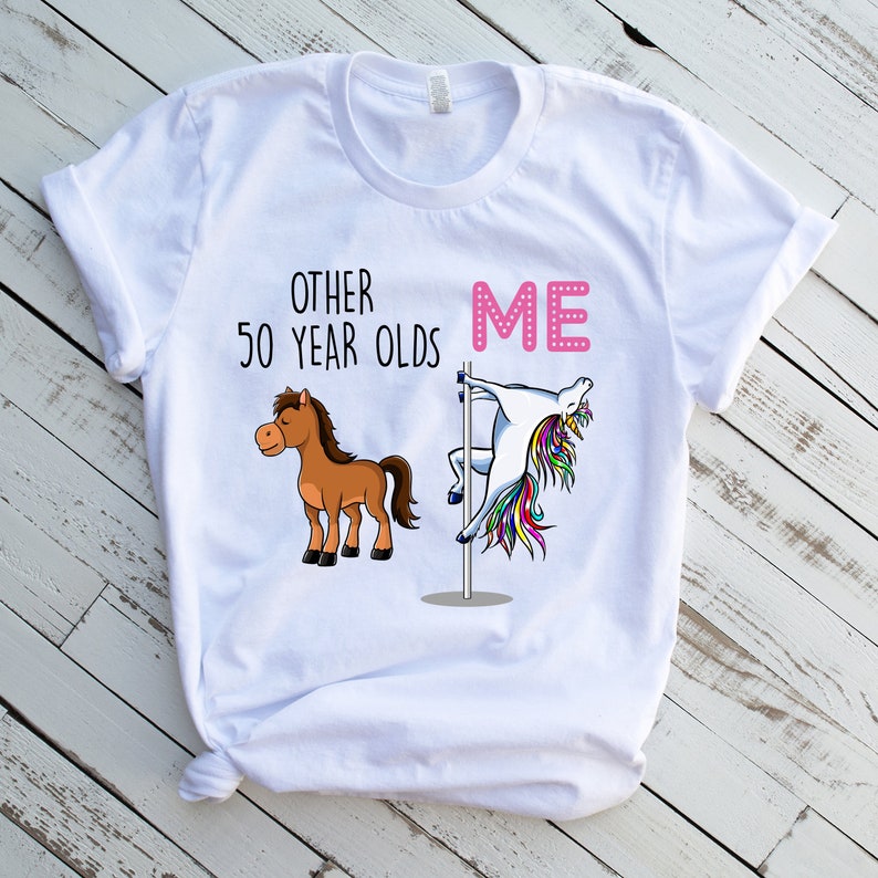 Pode incluir: Camiseta branca com um gr&aacute;fico de um cavalo e um unic&oacute;rnio. O texto "Other 50 year olds" est&aacute; acima do cavalo e o texto "Me" est&aacute; acima do unic&oacute;rnio. O unic&oacute;rnio est&aacute; em um poste e est&aacute; em uma pose de dan&ccedil;a.