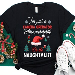 May include: Black t-shirt with a Christmas-themed design. The text reads "I'm just a camera operator whose permanently on the naughty list". The design includes a Santa hat, Christmas trees, and snowflakes.