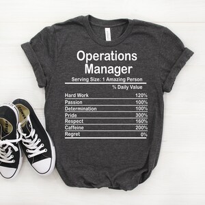 May include: A dark gray t-shirt with white text that reads "Operations Manager" and a nutrition facts label listing the percentage of daily value for hard work, passion, determination, pride, respect, caffeine, and regret.