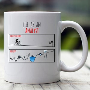 May include: White ceramic coffee mug with a black and white graphic depicting the expectations versus reality of being an analyst. The graphic shows a cyclist riding smoothly on a flat road in the expectations section and a cyclist struggling to climb a steep hill with obstacles in the reality section. The text "Life as an Analyst" is printed in red at the top of the mug.
