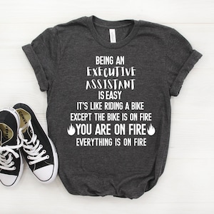 May include: A dark grey t-shirt with white text that reads "Being an Executive Assistant is easy. It's like riding a bike except the bike is on fire. You are on fire. Everything is on fire." There are two flame emojis on either side of the text "You are on fire."