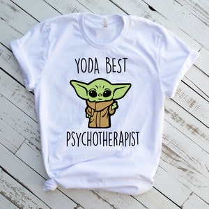 May include: White t-shirt with a green cartoon character and the text "Yoda Best Psychotherapist".