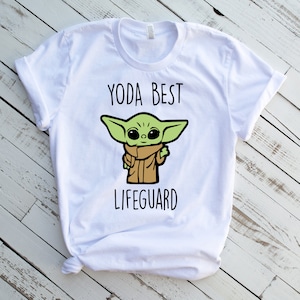 May include: White t-shirt with a green cartoon character wearing a brown coat and the text "Yoda Best Lifeguard".