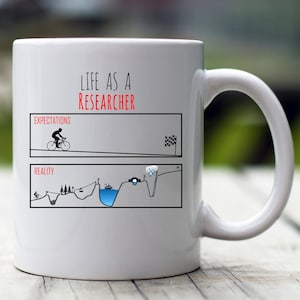 May include: White ceramic mug with a black and white illustration of a cyclist riding uphill. The text "Life as a Researcher" is printed in red on the mug. The illustration shows the cyclist's journey from expectations to reality, with obstacles and challenges along the way.