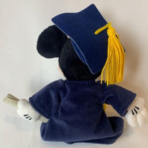 May include: A plush Mickey Mouse figure dressed in a navy blue graduation gown and cap. The cap has a yellow tassel. Mickey is holding a rolled-up diploma in his white-gloved hands. The figure is set against a white background.