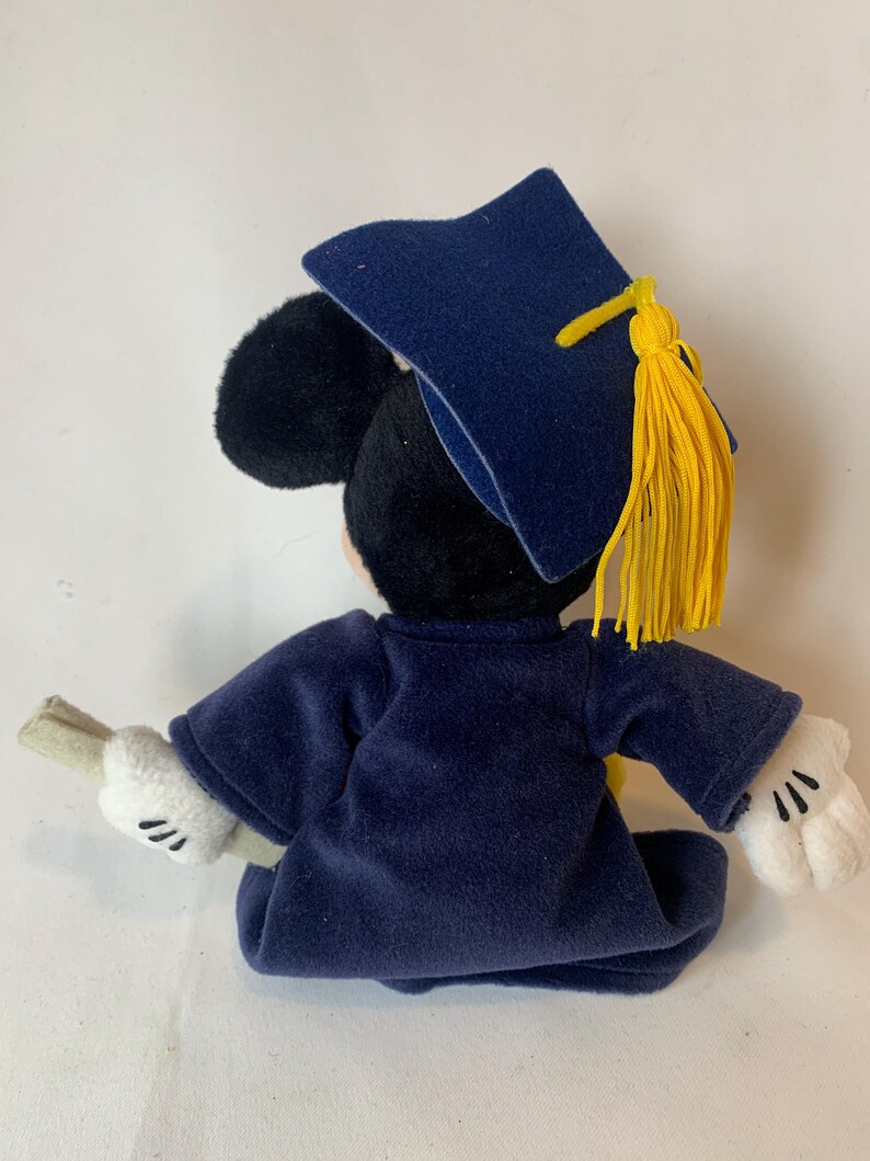May include: A plush Mickey Mouse figure dressed in a navy blue graduation gown and cap. The cap has a yellow tassel. Mickey is holding a rolled-up diploma in his white-gloved hands. The figure is set against a white background.