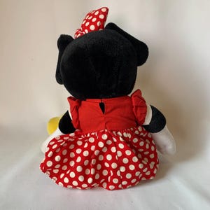 May include: A plush Minnie Mouse doll, viewed from the back, with a black head and ears, a red dress with white polka dots, and a red bow with white polka dots. The doll has white gloves and yellow shoes.