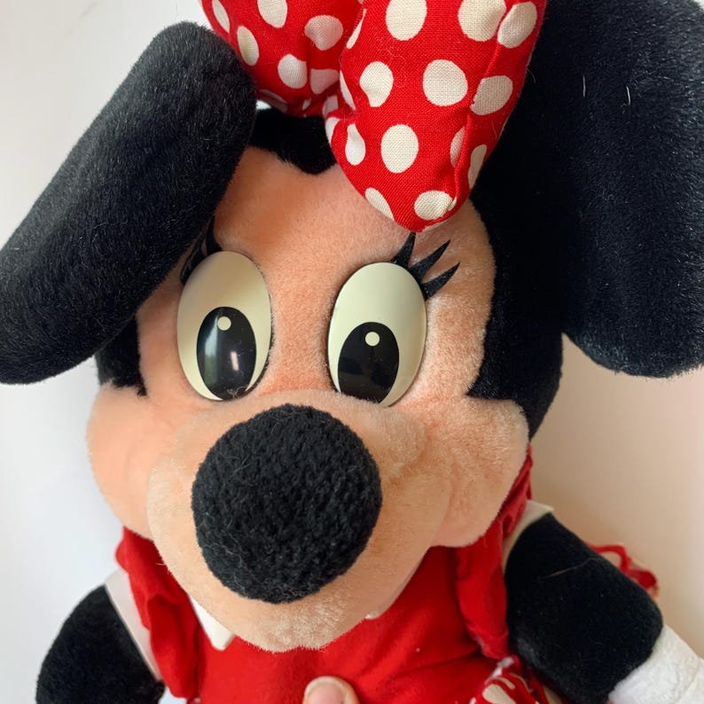 May include: A plush Minnie Mouse doll with a red dress, a white collar, and a red bow with white polka dots. The doll has black ears, a black nose, and large, expressive eyes. The doll is a classic Disney character.