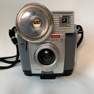 May include: Vintage Kodak Flashmite 20 camera with a silver lens and flash, gray body, and black strap. The camera has the Kodak logo and the words "Flashmite 20 Camera" printed on the front. The camera has a color setting of 13, 14, and 15.
