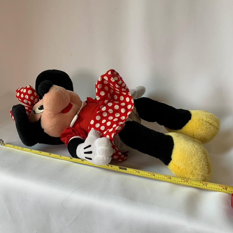 May include: A plush Minnie Mouse doll, lying on its side. The doll features a red dress with white polka dots, a matching bow, and black ears. The doll's feet are yellow, and it has white gloves. A yellow measuring tape is next to the doll.