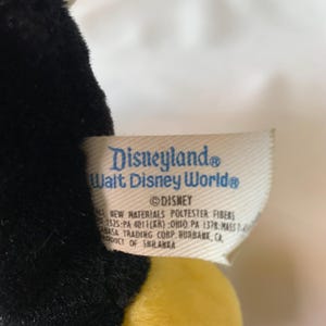May include: A close-up of a tag from a Disney plush toy. The tag is white with blue text that reads "Disneyland" and "Walt Disney World". The Disney logo is also visible. The tag also includes information about the materials and manufacturer.