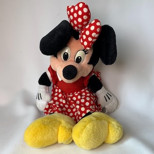 May include: A plush Minnie Mouse doll, featuring a red dress with white polka dots and a matching bow. The doll has black ears, a black nose, and yellow shoes. The doll is sitting down and has white gloves.