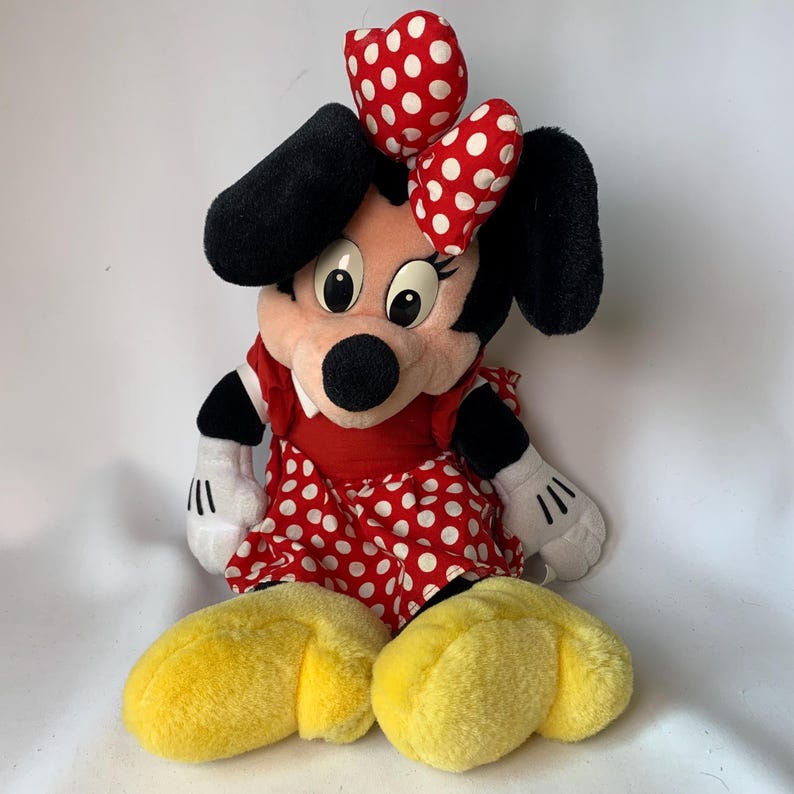 May include: A plush Minnie Mouse doll, featuring a red dress with white polka dots and a matching bow. The doll has black ears, a black nose, and yellow shoes. The doll is sitting down and has white gloves.