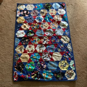 May include: A handmade quilt with a patchwork design. The quilt features a variety of patterned fabrics in a hexagonal pattern, with red and blue triangles. The fabrics include various prints, such as animals, flowers, and geometric shapes. The quilt is bordered with a solid blue fabric.