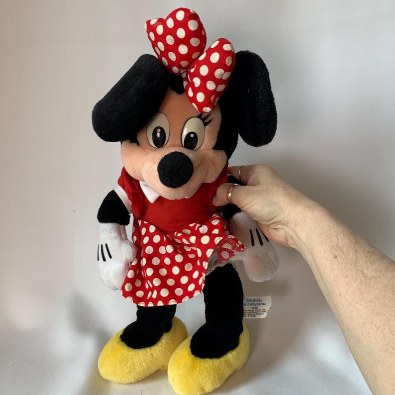 May include: A Minnie Mouse plush doll, featuring a red dress with white polka dots, a matching bow, and yellow shoes. The doll has black ears, a black nose, and white gloves. The doll is a classic Disney character.