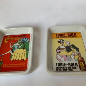 May include: Two white ceramic rectangular trays with colorful vintage advertisements. The left tray features a woman holding a glass of champagne with the text "De la Germanie Champagne". The right tray features a couple sitting at a table with the text "Tonie-Kola Vin Aperitif a la Kola".