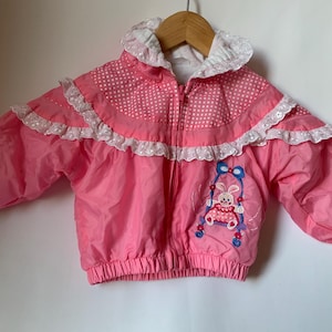 May include: A pink and white jacket with a white lace trim and a bunny embroidered on the front. The jacket has a zipper closure and a white hood.