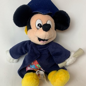 May include: A plush Mickey Mouse doll wearing a navy blue graduation cap and gown. The doll has black ears, a tan face, and yellow feet. It holds a rolled diploma. A tag with the Disney logo is attached.