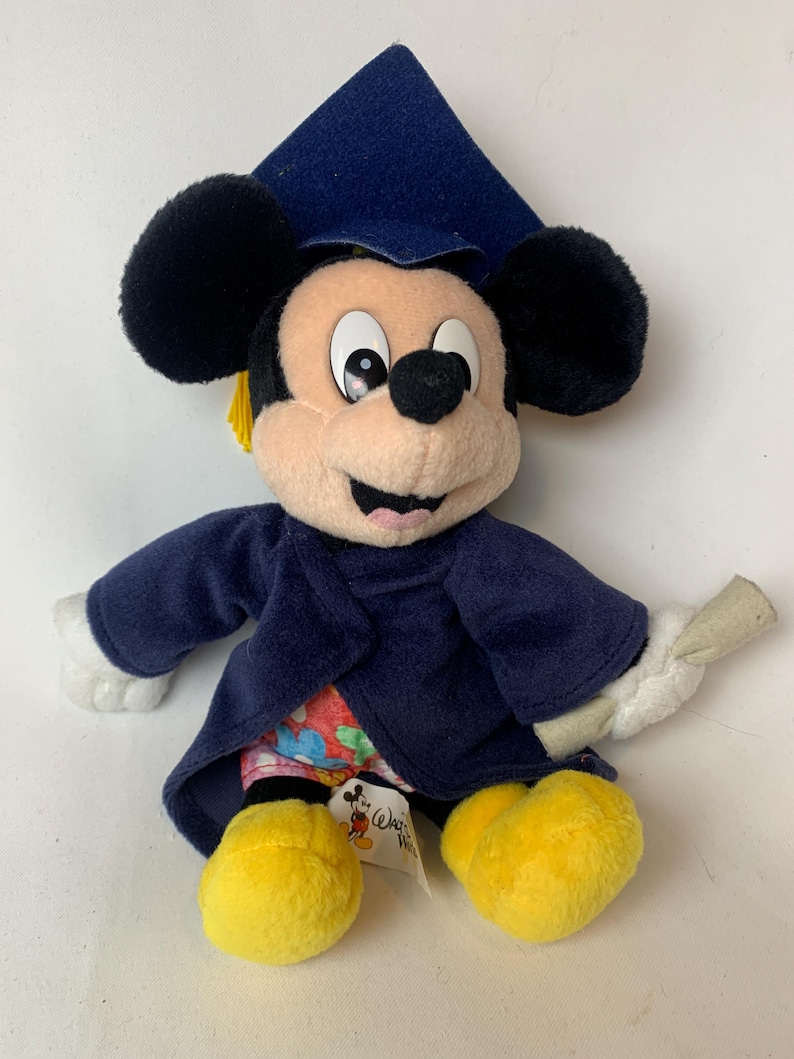 May include: A plush Mickey Mouse doll wearing a navy blue graduation cap and gown. The doll has black ears, a tan face, and yellow feet. It holds a rolled diploma. A tag with the Disney logo is attached.