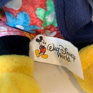 May include: A close-up of a Mickey Mouse plush toy. The toy has yellow gloves, black shorts, and a blue jacket. The interior lining features a floral pattern. A tag with the Walt Disney World logo and Mickey Mouse is visible.