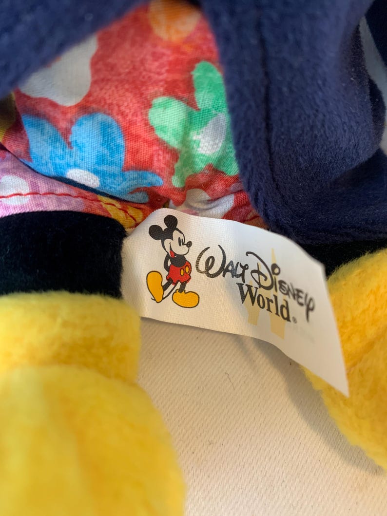 May include: A close-up of a Mickey Mouse plush toy. The toy has yellow gloves, black shorts, and a blue jacket. The interior lining features a floral pattern. A tag with the Walt Disney World logo and Mickey Mouse is visible.