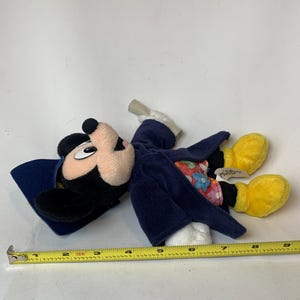May include: A Mickey Mouse plush toy wearing a blue coat and hat, with yellow gloves and shoes. The toy has a beige face and black ears. A measuring tape is next to the toy.