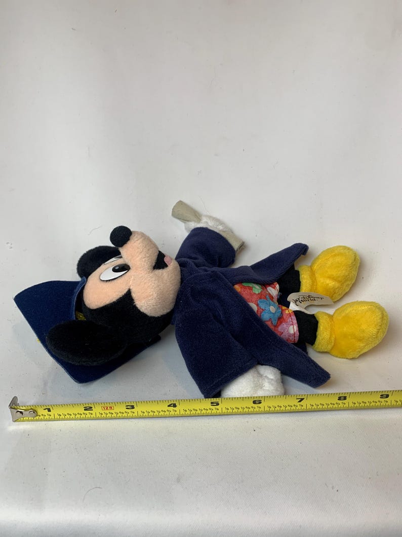 May include: A Mickey Mouse plush toy wearing a blue coat and hat, with yellow gloves and shoes. The toy has a beige face and black ears. A measuring tape is next to the toy.