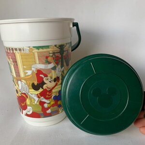 May include: A white plastic souvenir cup with a green handle and lid. The cup features a colorful graphic of Disney characters, including Minnie Mouse, in a parade setting. The green lid has a Mickey Mouse silhouette.
