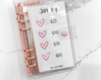 May include: A clear plastic binder with a pink heart design and the text "Currently" and "Jani ary" on the front. The binder has a pink ring mechanism and a pink heart sticker on the front. The text "Ada Layne Budgets" is printed on the bottom of the page.