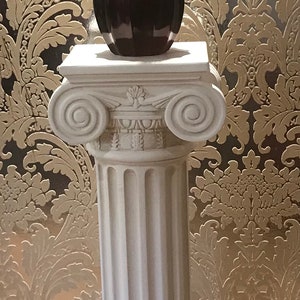 Op de afbeelding: Een witte decoratieve zuil ondersteunt een rood-bruine vaas met gedroogde takken. De zuil heeft een klassiek ontwerp. De vaas heeft een unieke vorm. De zuil is ongeveer 100 cm hoog.