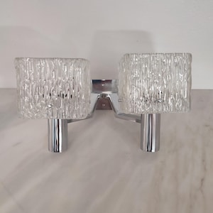 May include: A dual-light wall sconce featuring two square, textured clear glass shades mounted on a polished chrome metal frame. The fixture has two cylindrical chrome sockets extending from a central rectangular base plate.