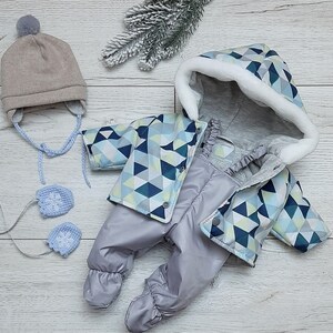 Könnte beinhalten: Ein grauer und blauer Baby-Schneeanzug mit geometrischem Muster und einer weißen, pelzgefütterten Kapuze. Der Schneeanzug hat einen Reißverschluss und eine passende Mütze.