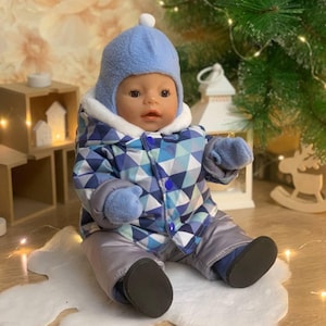 May include: A baby doll wearing a blue winter hat with a white pom-pom, a blue and white geometric patterned jacket, gray pants, and blue mittens. The doll is sitting on a white, cloud-shaped rug.