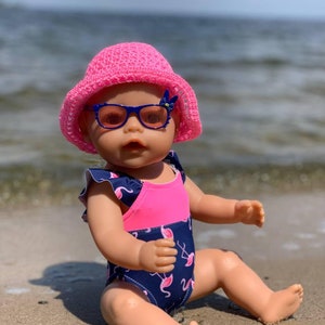Könnte beinhalten: Ein rosa-blauer Badeanzug mit Flamingomuster, getragen von einer Puppe mit rosa Sonnenbrille und einem rosa Häkelhut. Die Puppe sitzt auf einem Sandstrand.