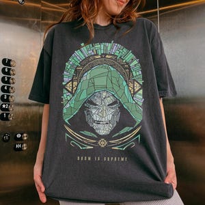 Può includere: Una t-shirt a maniche corte grigio antracite con la grafica di una figura mascherata con cappuccio verde in stile vetrata. Il design include intricate linee geometriche e il testo DOOM IS SUPREME stampato con un carattere color oro.