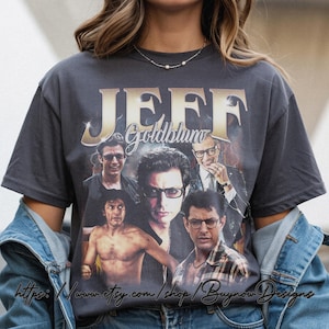 May include: Dark gray t-shirt featuring a collage of Jeff Goldblum images with the name "JEFF Goldblum" in gold lettering. The design includes various photos of the actor, creating a vintage-style graphic.