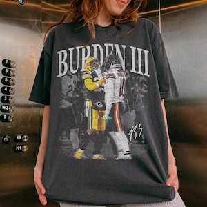 May include: A charcoal grey short-sleeve t-shirt featuring a graphic print of two football players in team uniforms. The text BURDEN III is printed in large white letters above the players, and the number 13 is written in white at the bottom right.